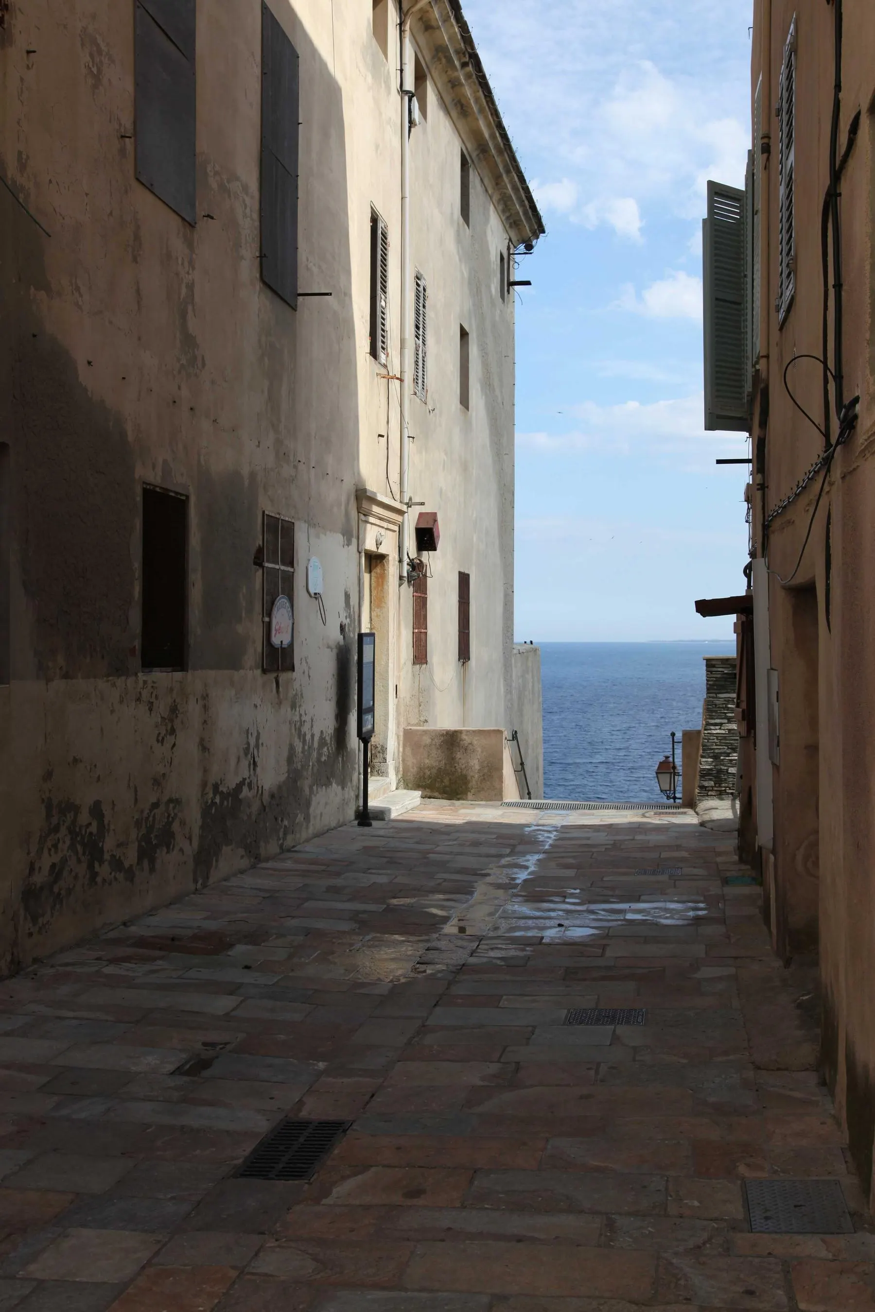 A narrow stone alley between old buildings opening onto a view of the blue sea.