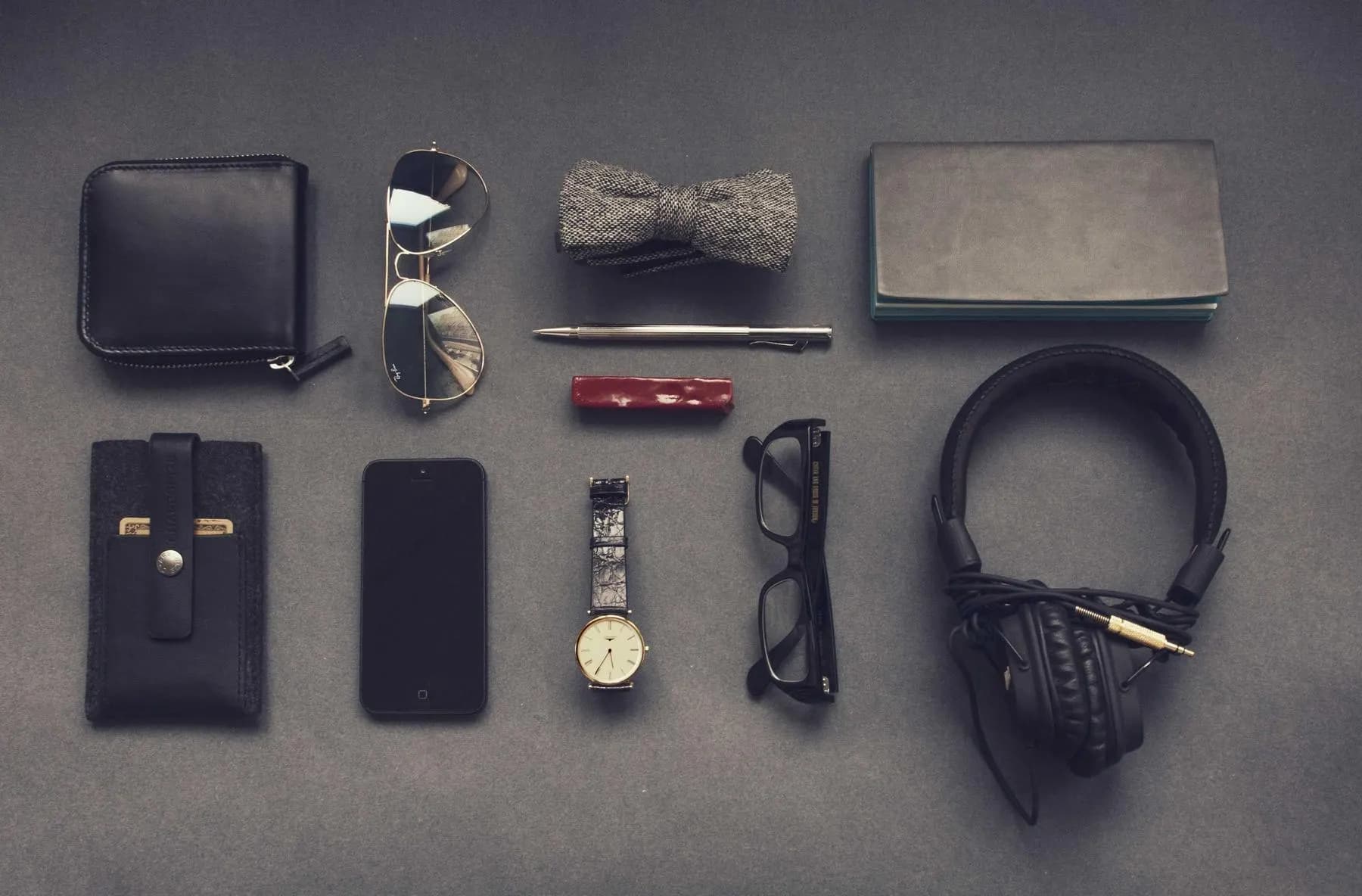 Everyday carry laid out on grey felt: a wallet, sunglasses, a bow tie, a notebook, a phone, a watch, glasses and headphones.