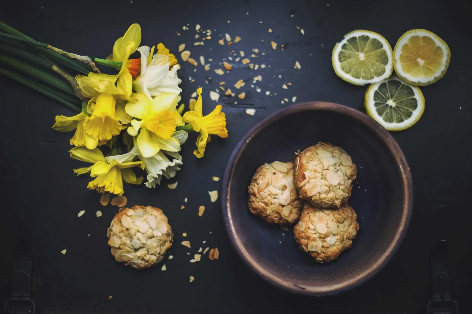 Yellow daffodils, lemon slices and a bowl of round biscuits scattered across a dark table.