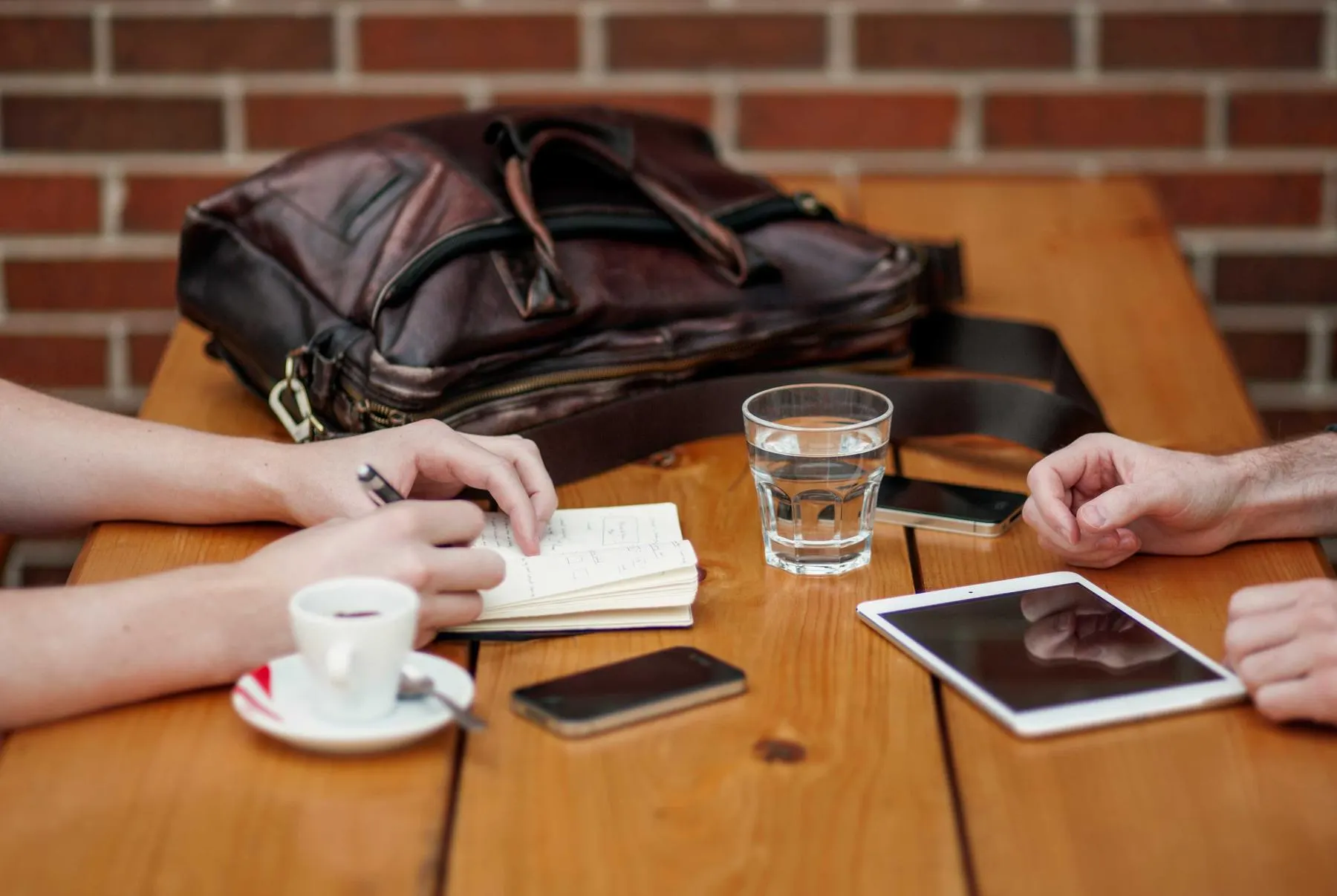 A brown leather bag on a café table between two people, a notebook, a glass of water and a cup of coffee.