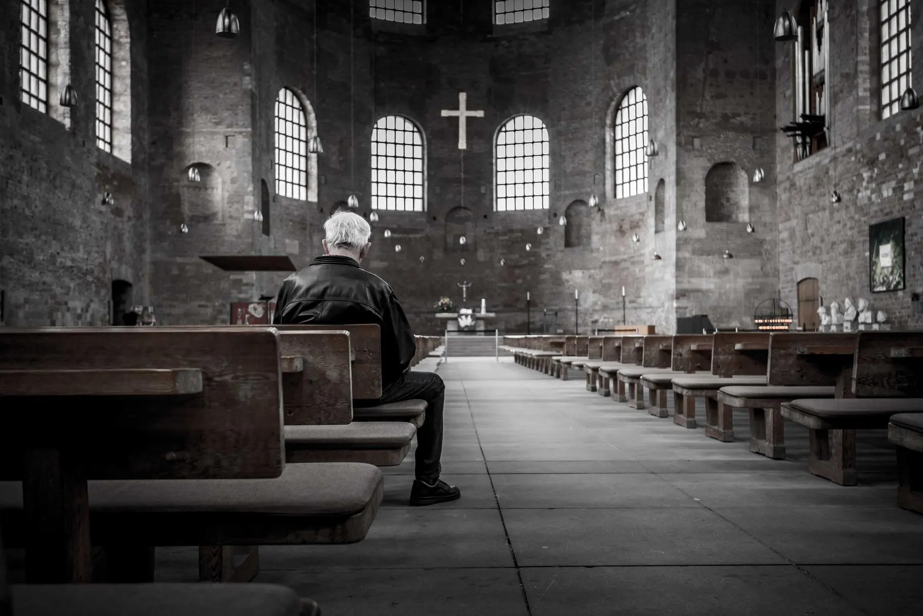 A man sitting alone on a wooden pew in a large stone hall with arched windows.