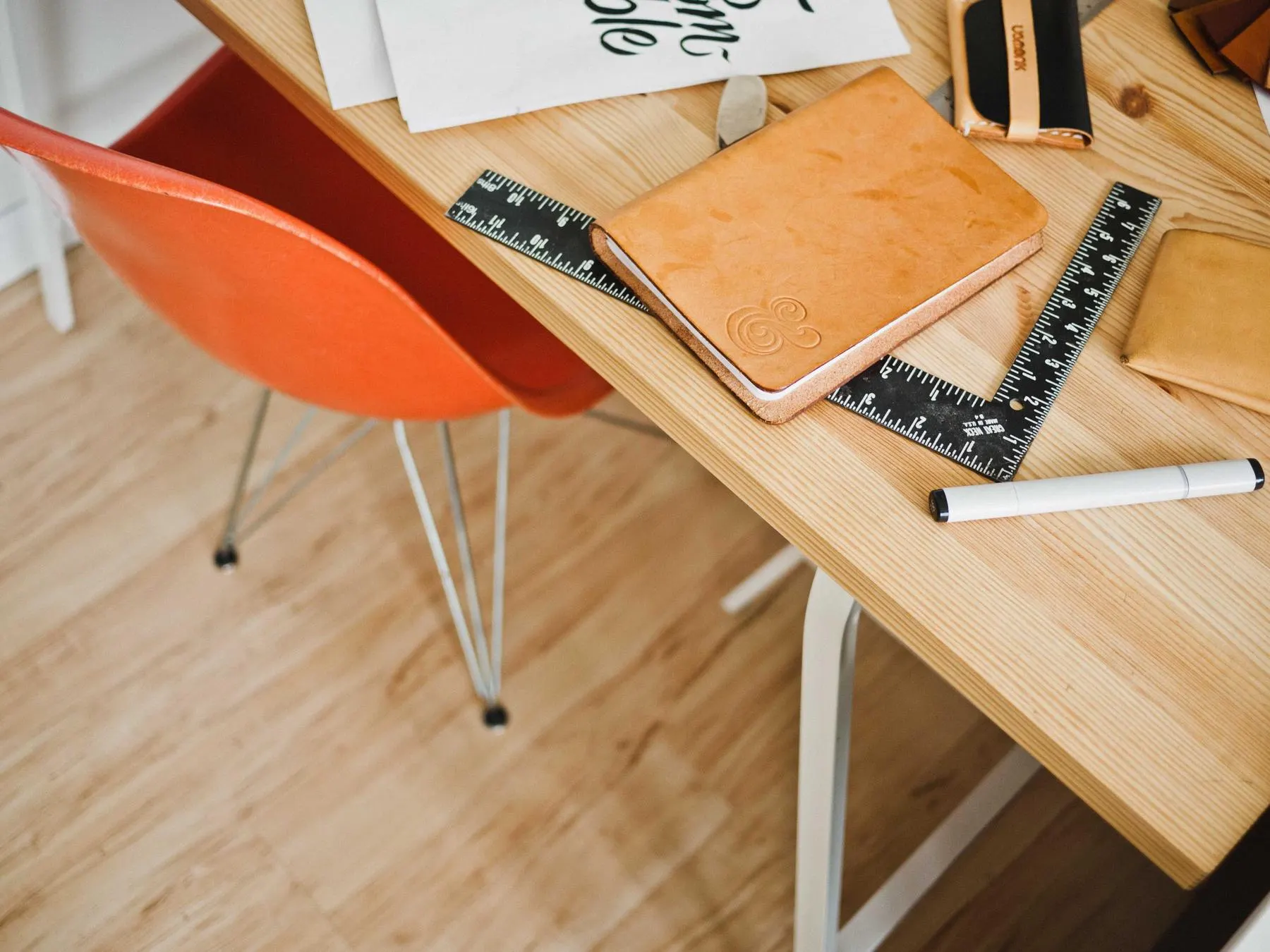 A wooden desk with a leather notebook, a metal ruler, a set square and pens beside an orange chair.