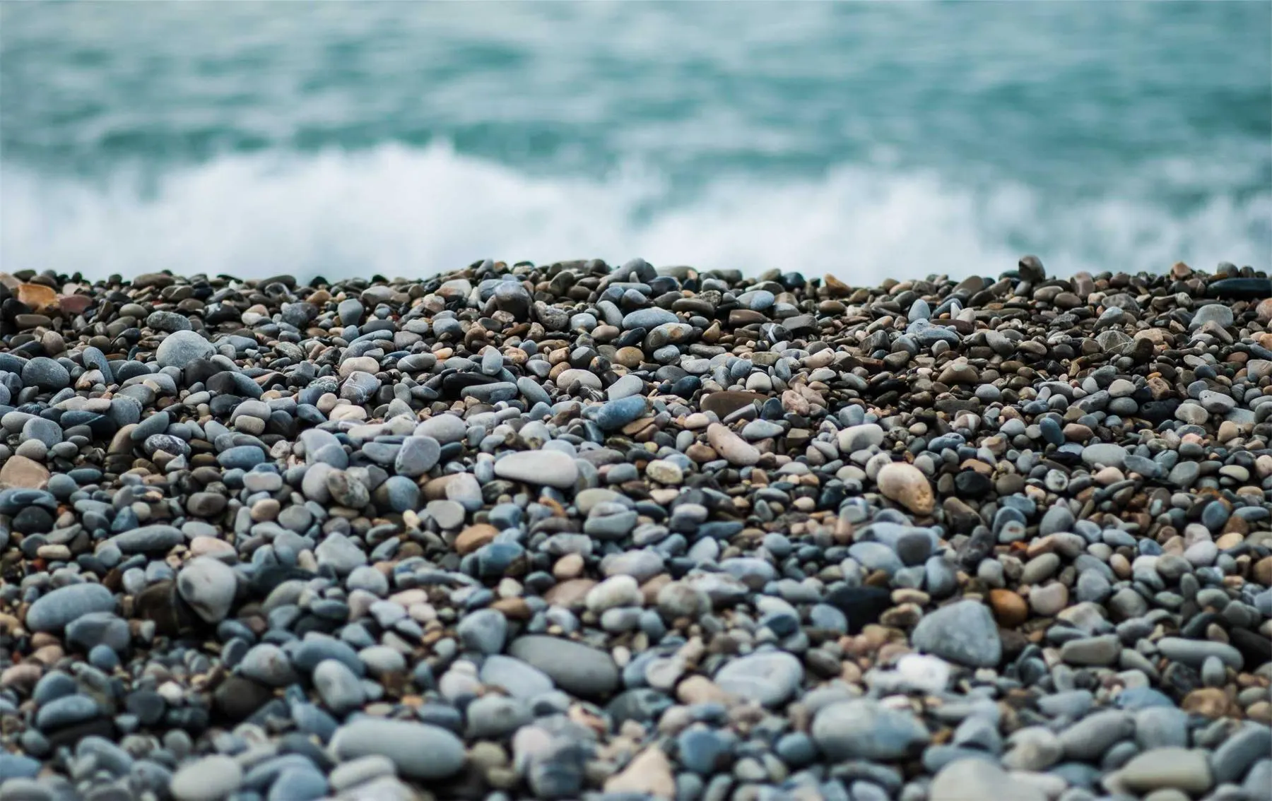 A beach of smooth grey and blue pebbles with a wave breaking softly behind them.