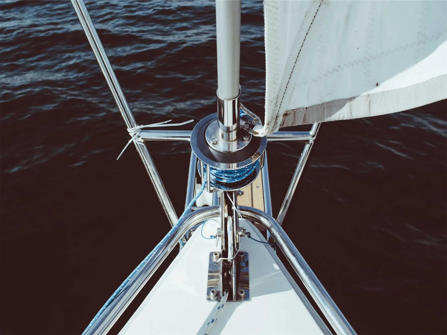 Looking down along the bow of a sailing boat, rigging and a white sail over dark water.