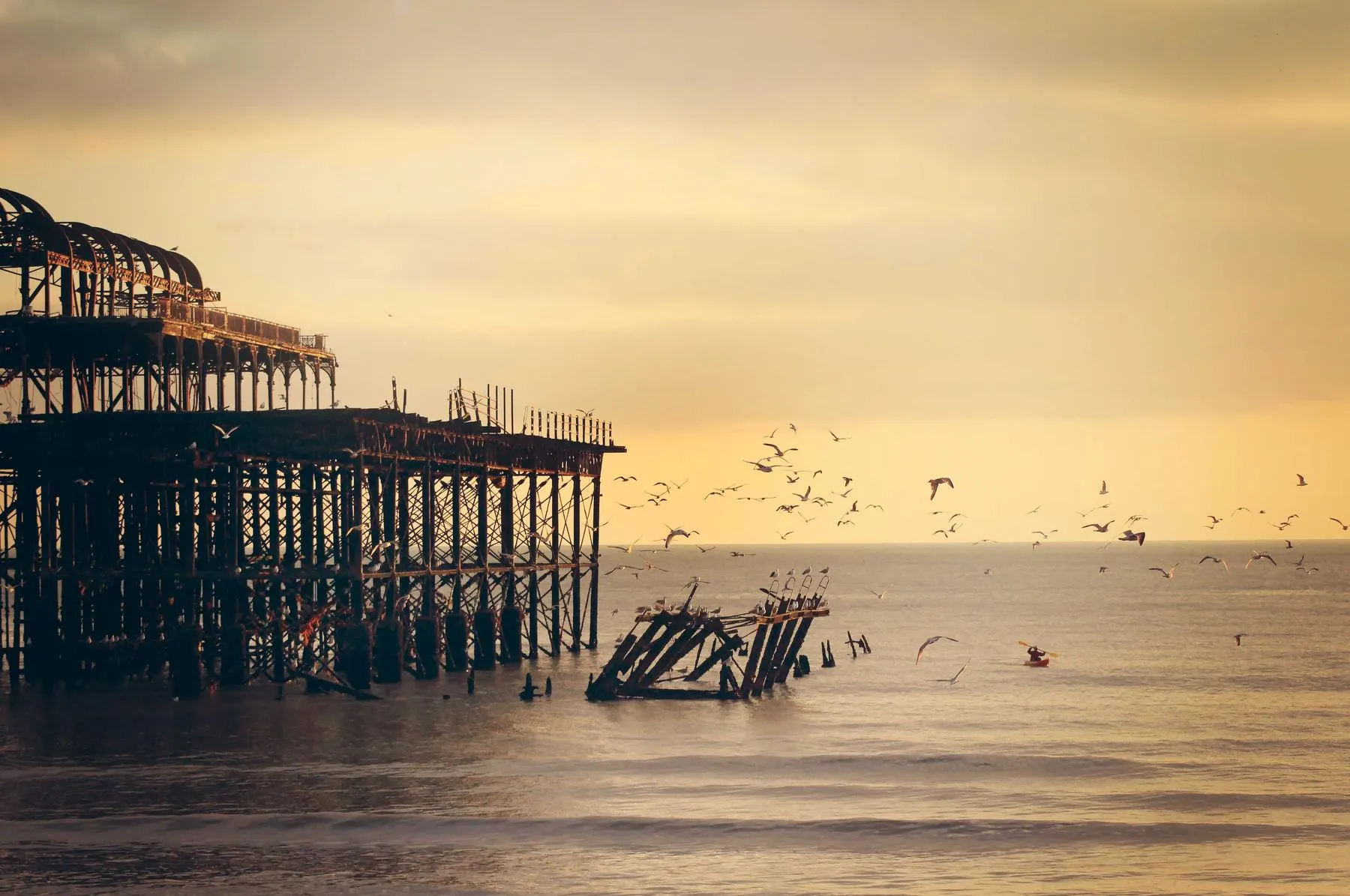 The iron skeleton of an old pier standing in the sea under a golden sky full of birds.