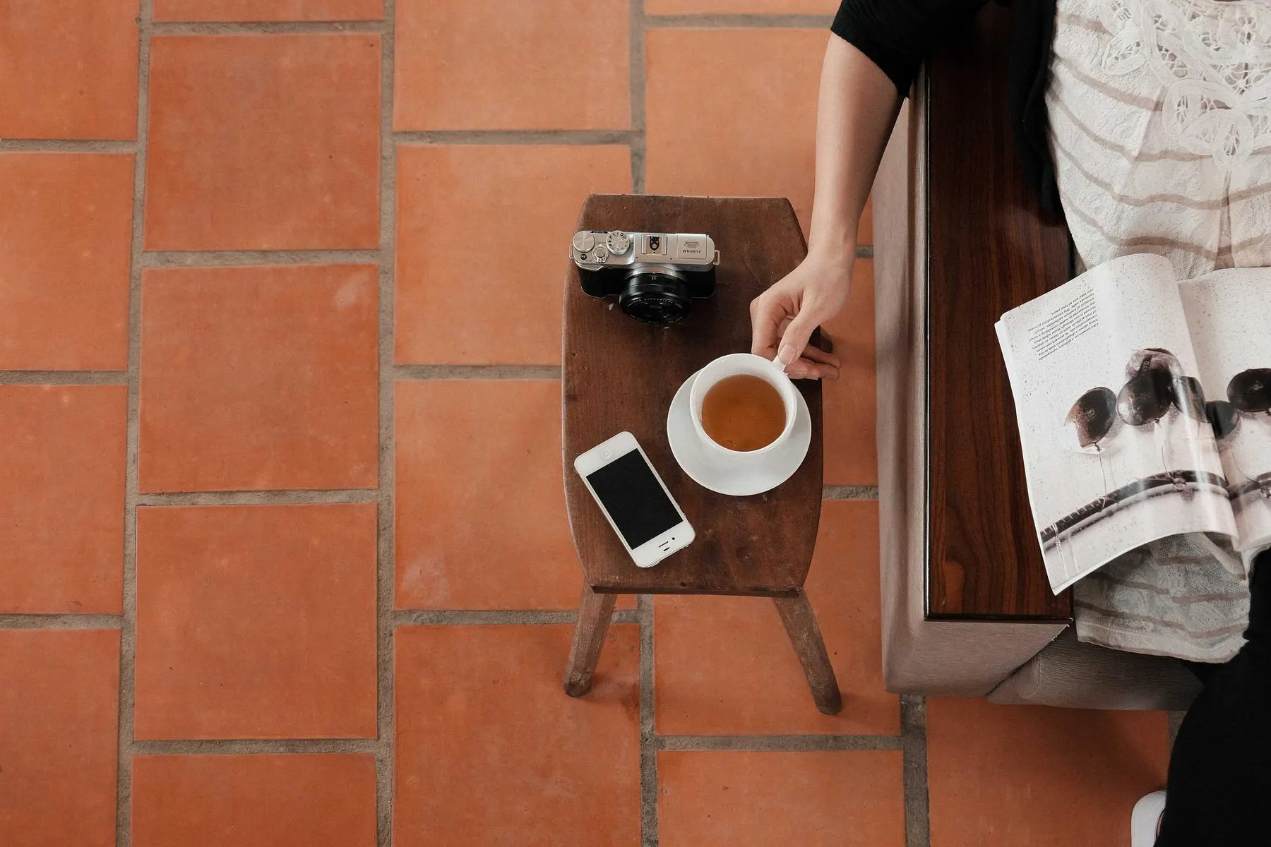Seen from above, a person reading a magazine beside a small table with a cup of tea, a phone and a camera on terracotta tiles.