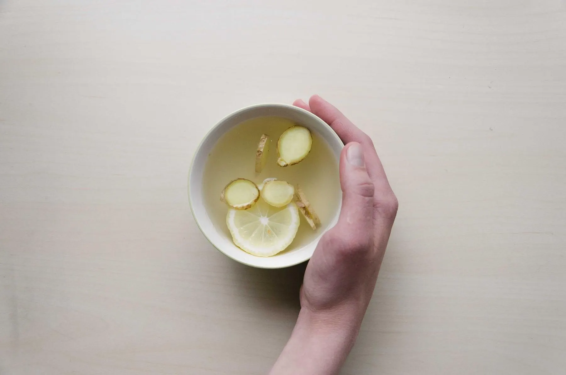 A hand resting around a white bowl of tea with slices of lemon and ginger on a pale table.