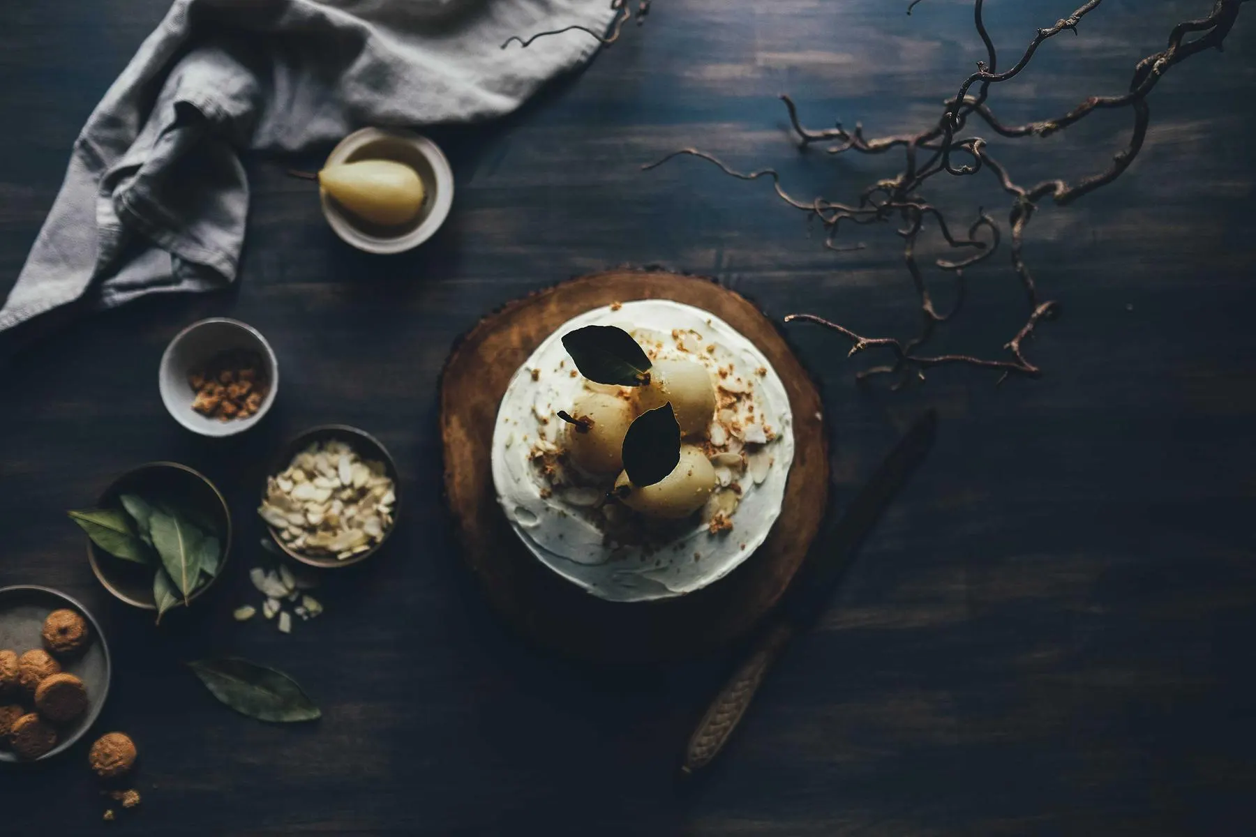 Seen from above, a dark wooden table set with a cake, small bowls of nuts and spices, a linen cloth and bare branches.
