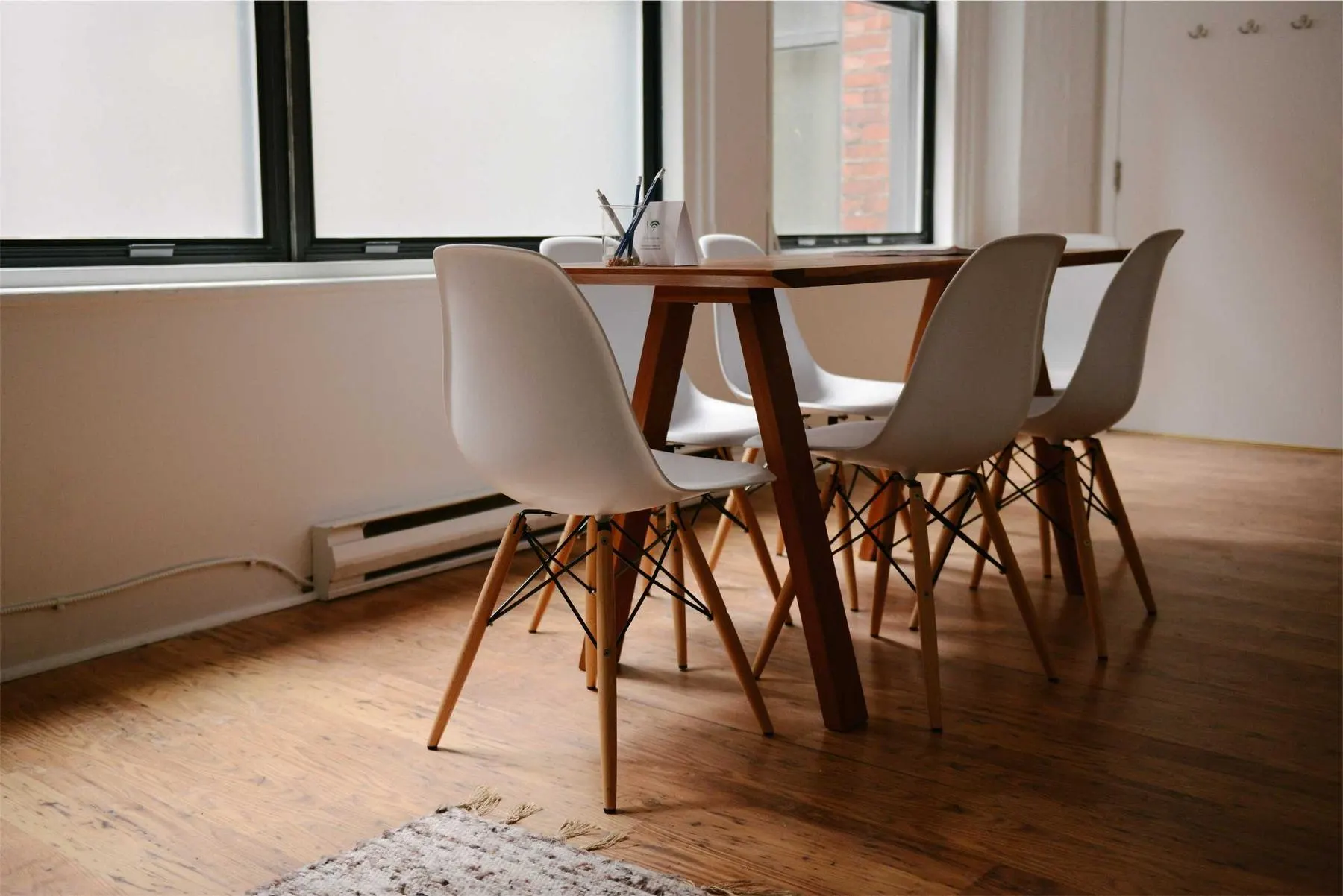 A bright, nearly empty dining room with a wooden table, white chairs and tall windows letting in soft daylight.